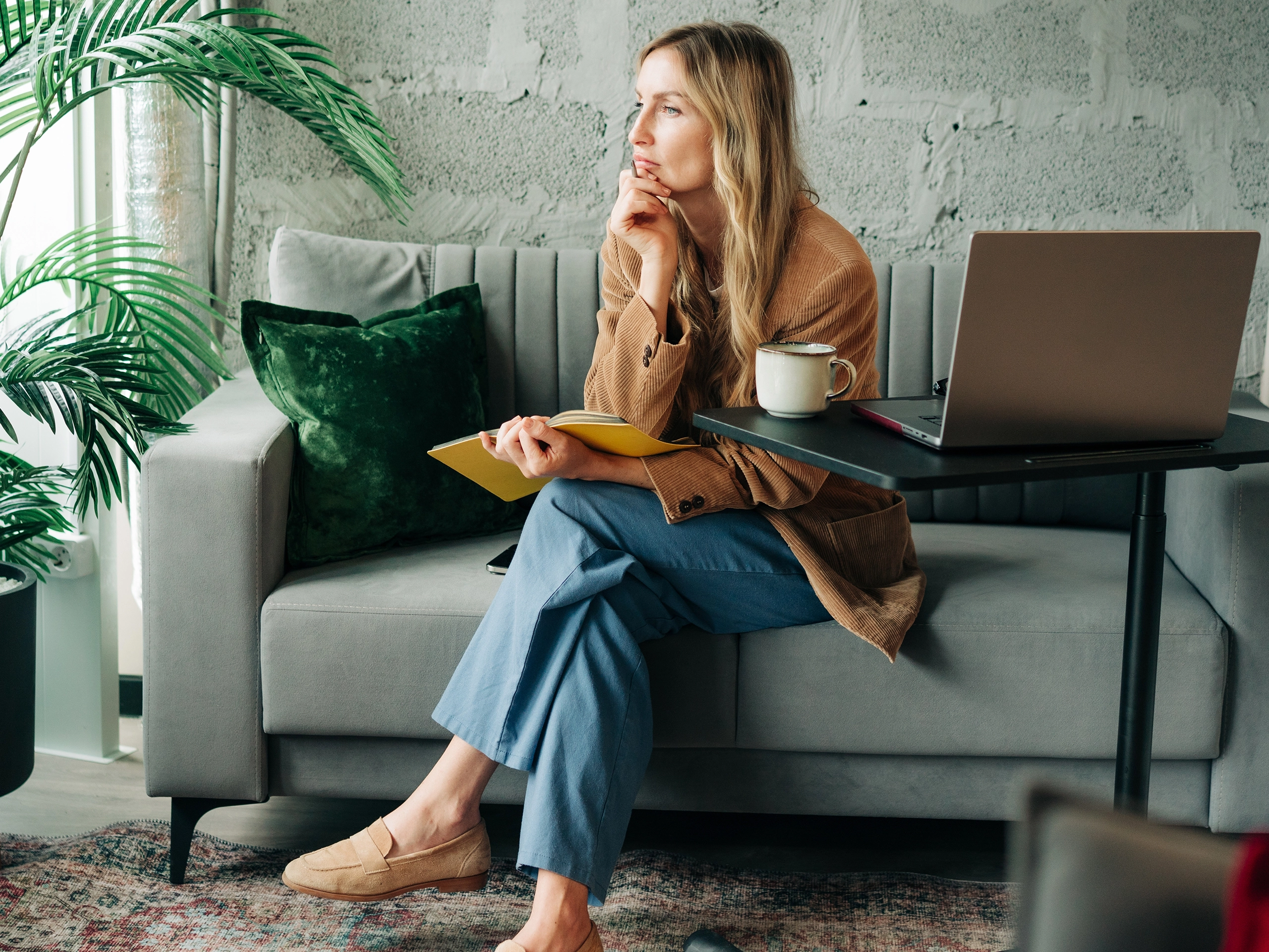 Woman working from home on laptop, thoughtful and relaxed on couch with notebook and coffee