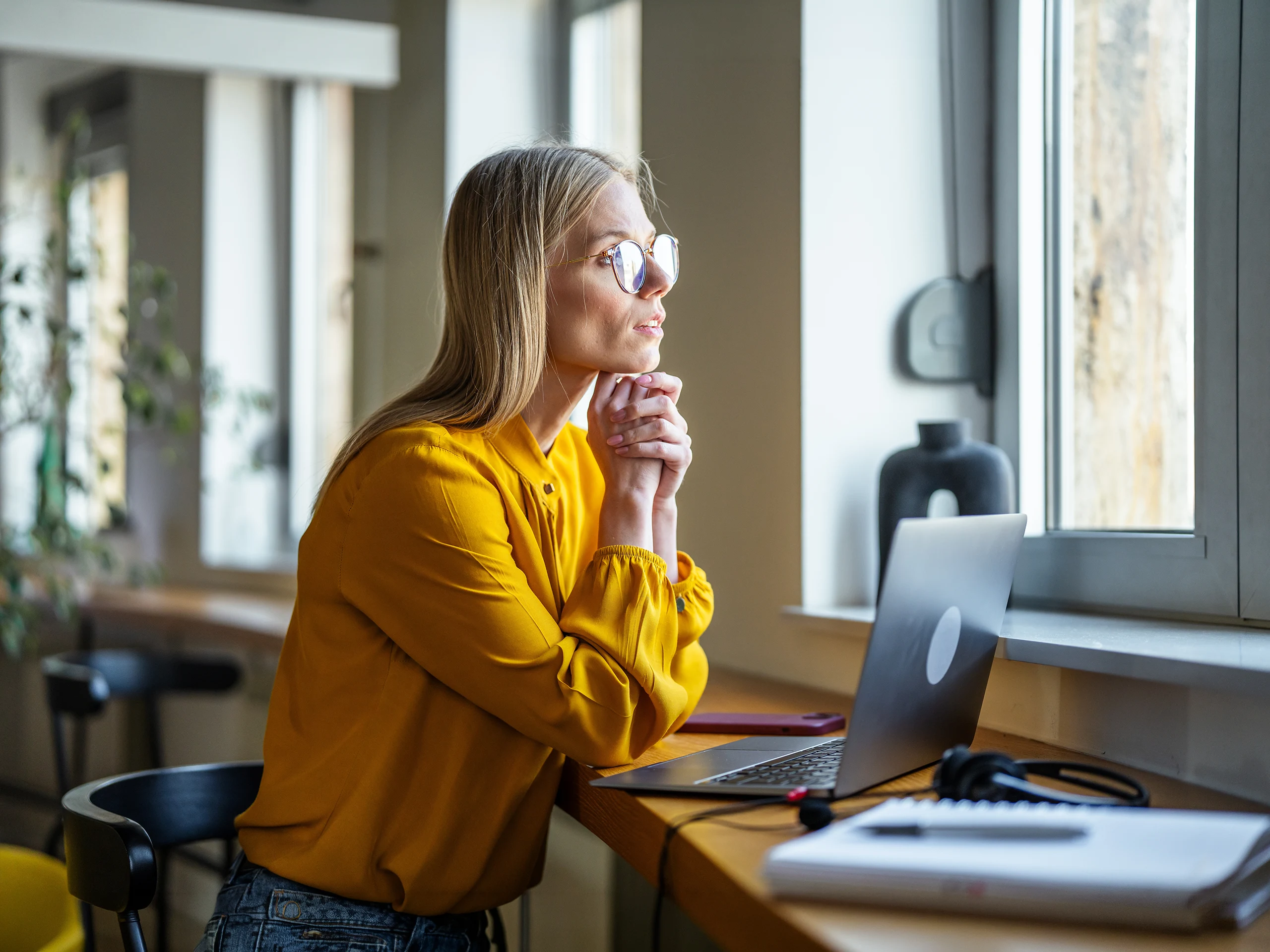 Young businesswoman with glasses and blonde hair thinking and looking out the window, planning strategies while working remotely with a laptop and headphones at her home office desk