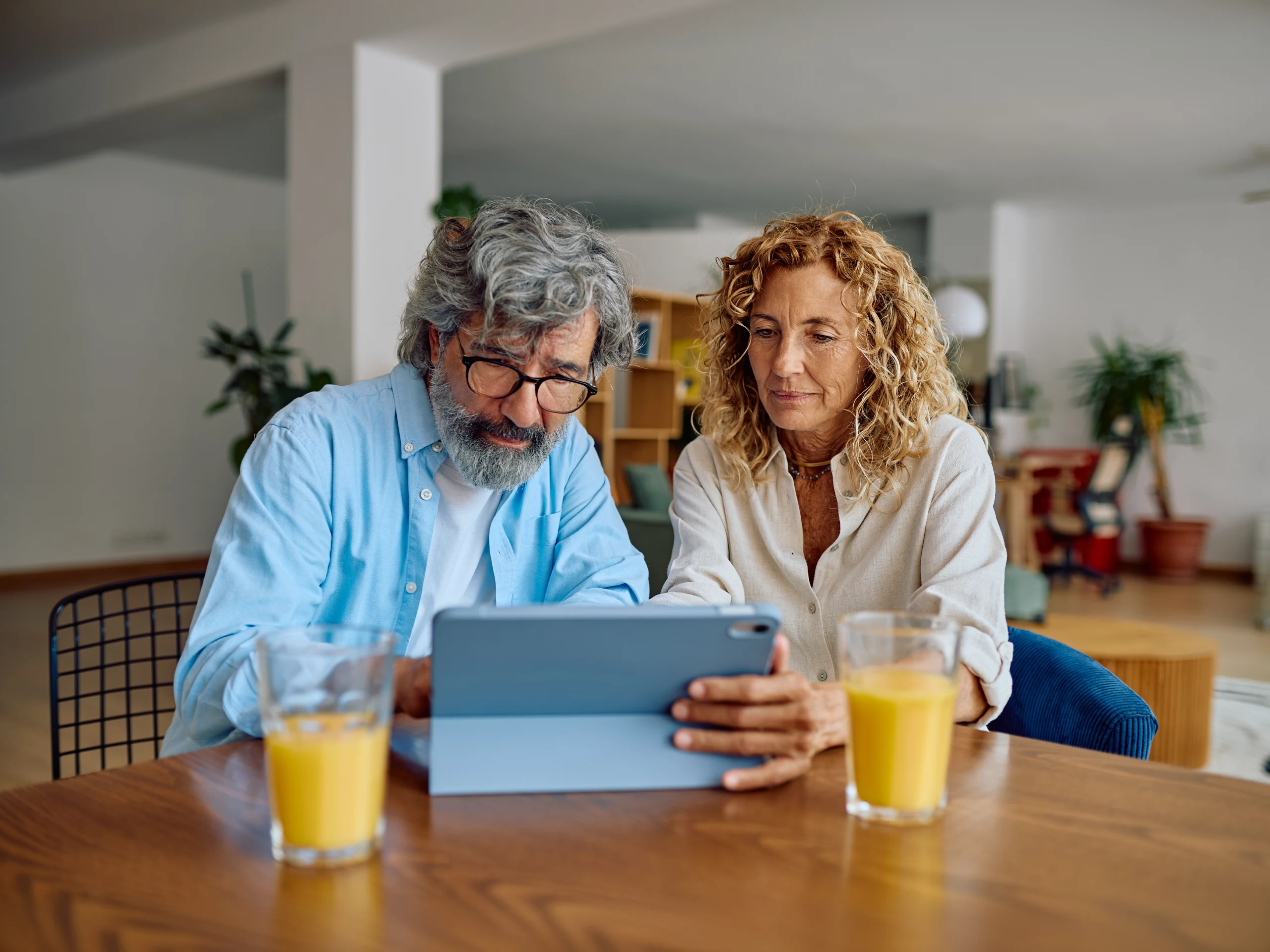 Senior couple using digital tablet and drinking orange juice while sitting at dining table in their modern apartment