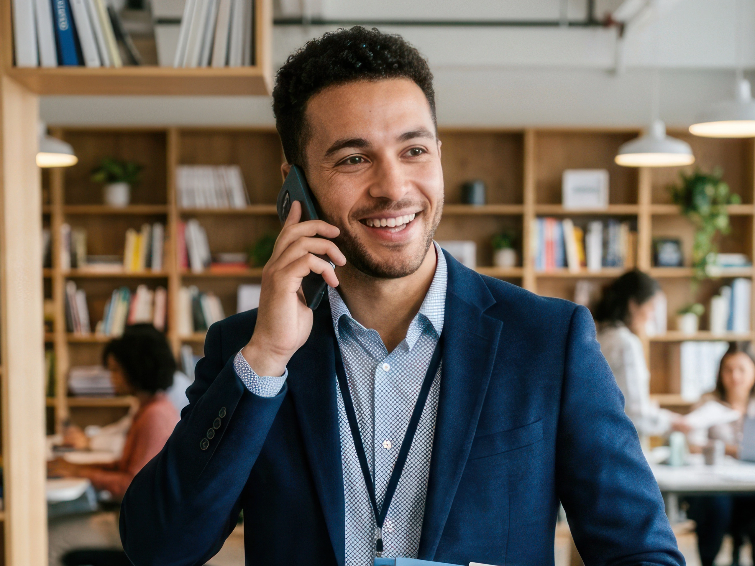 A smiling young man in a blue suit is talking on his phone while walking through a modern office. He is holding a stack of documents and appears to be engaged in a business conversation