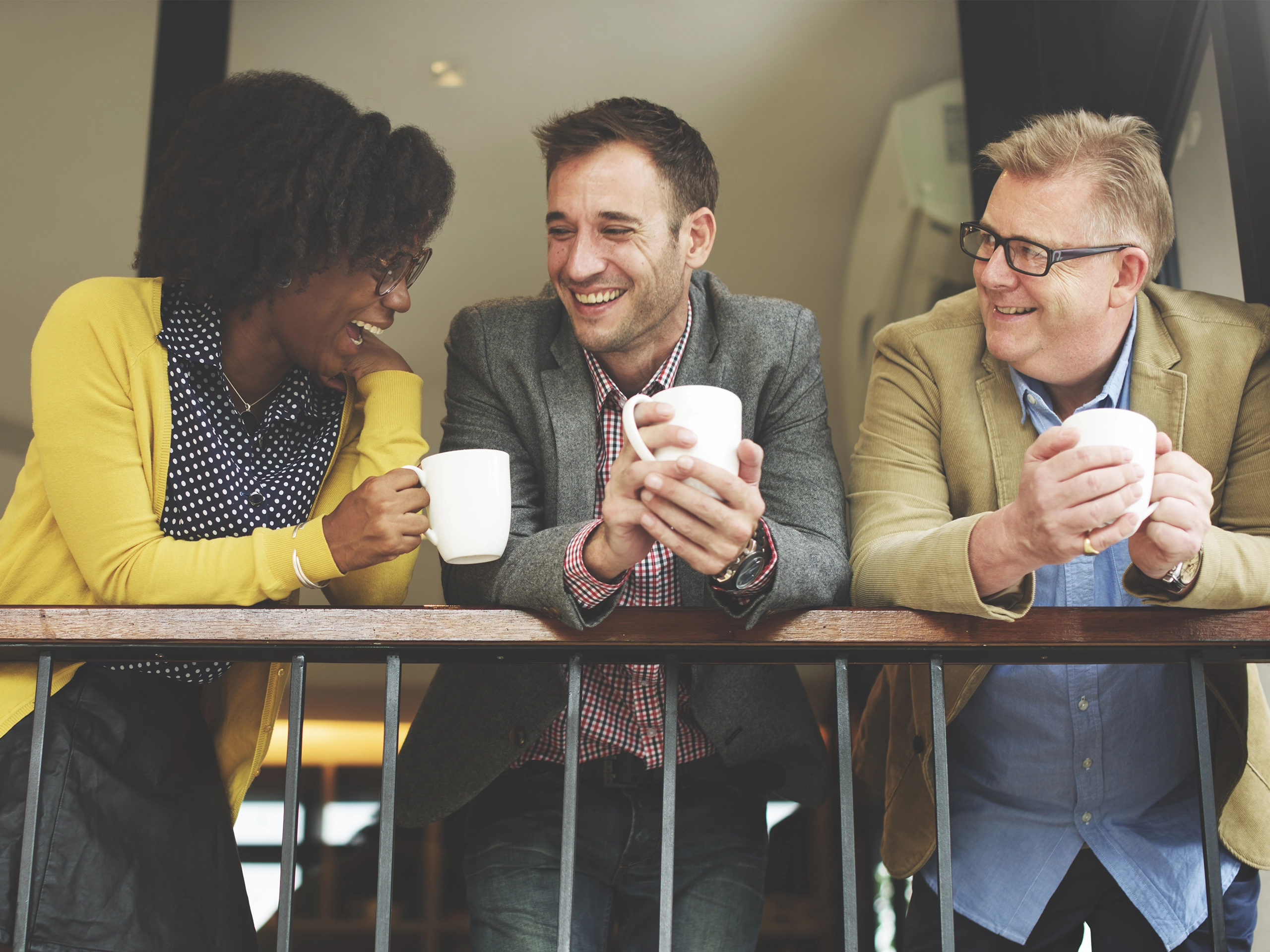 Colleagues chatting over coffee mugs leaning on balcony in workplace