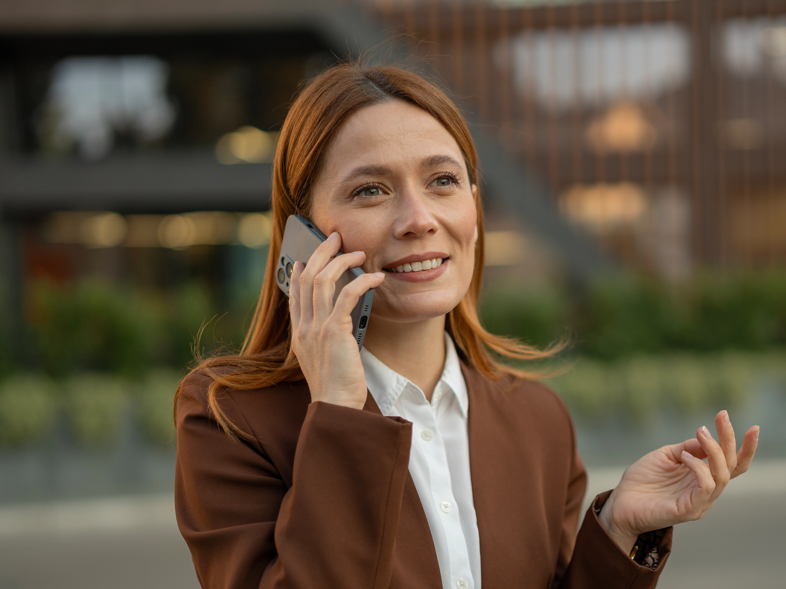 Professional young adult woman wearing a brown blazer and white shirt, happily communicating on a mobile phone outdoors in an urban setting, expressing confidence and connection