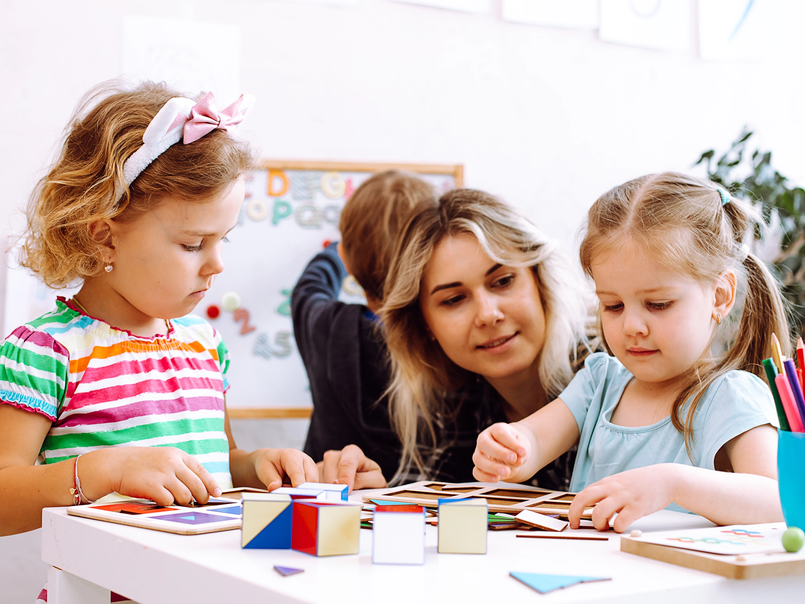 Little girls with interest fold colored cubes on desk in playroom of kindergarten. Young educator sitting with children at table on floor and watch play. Lesson in daycare centre