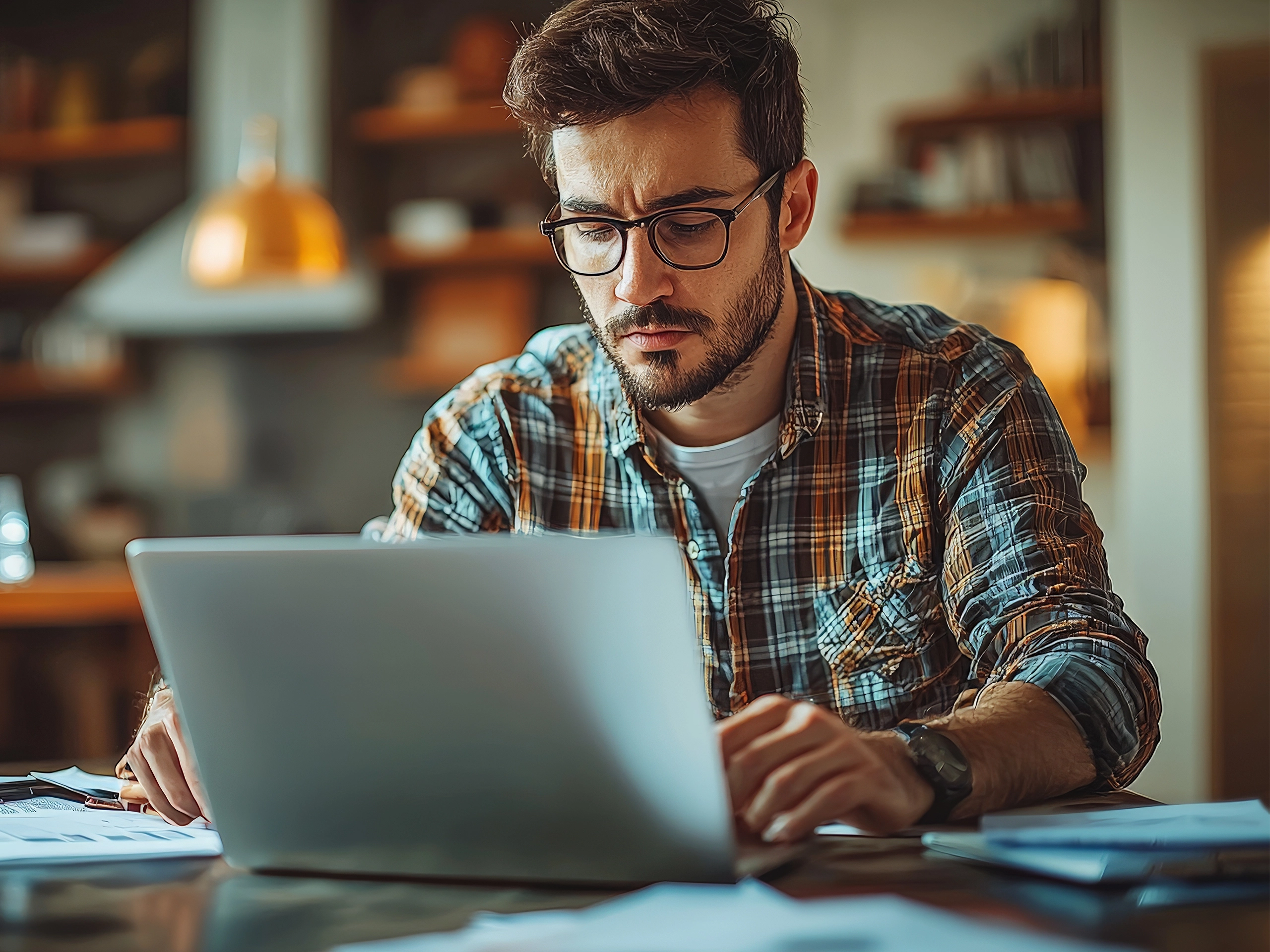 A focused man working on a laptop at a cozy home office with coffee and paperwork.