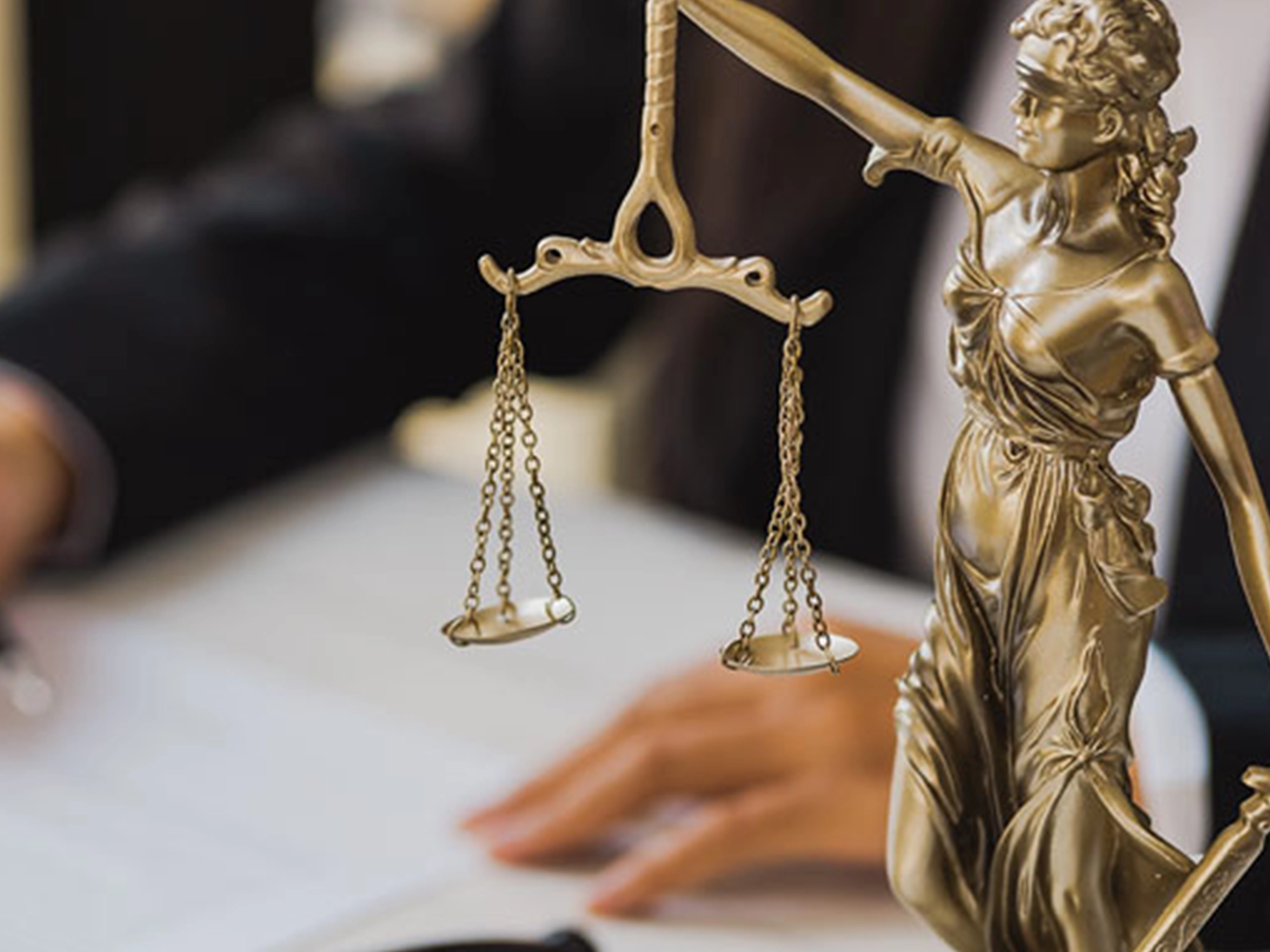 A close-up of a gold Lady Justice statue holding balanced scales, symbolising fairness and justice, set against a blurred office background with a desk and a person working.