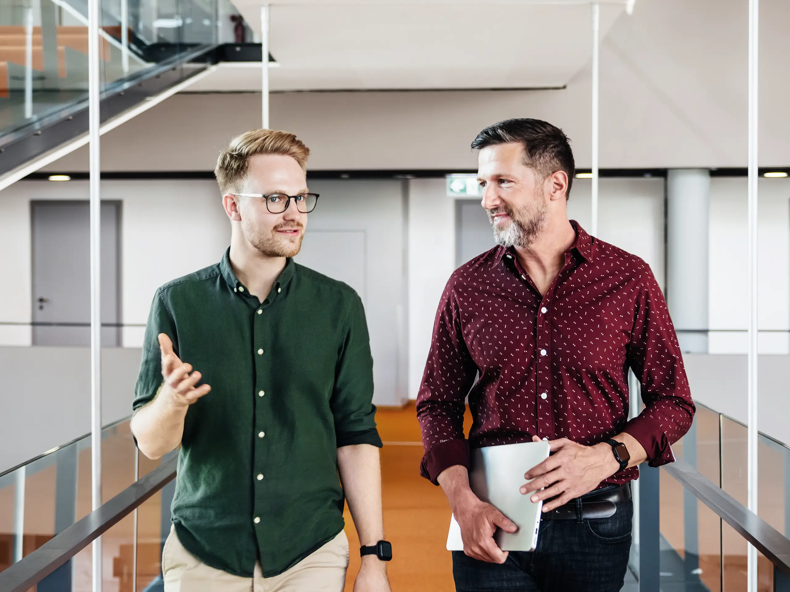 Two people walk through a modern office area while talking, with one person holding a tablet.