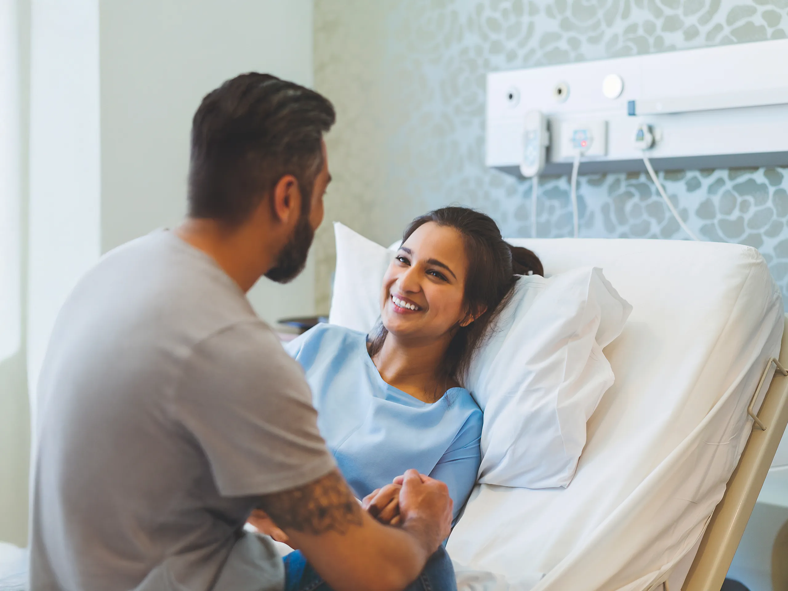 Smiling woman in a hospital bed holding a man’s hand.