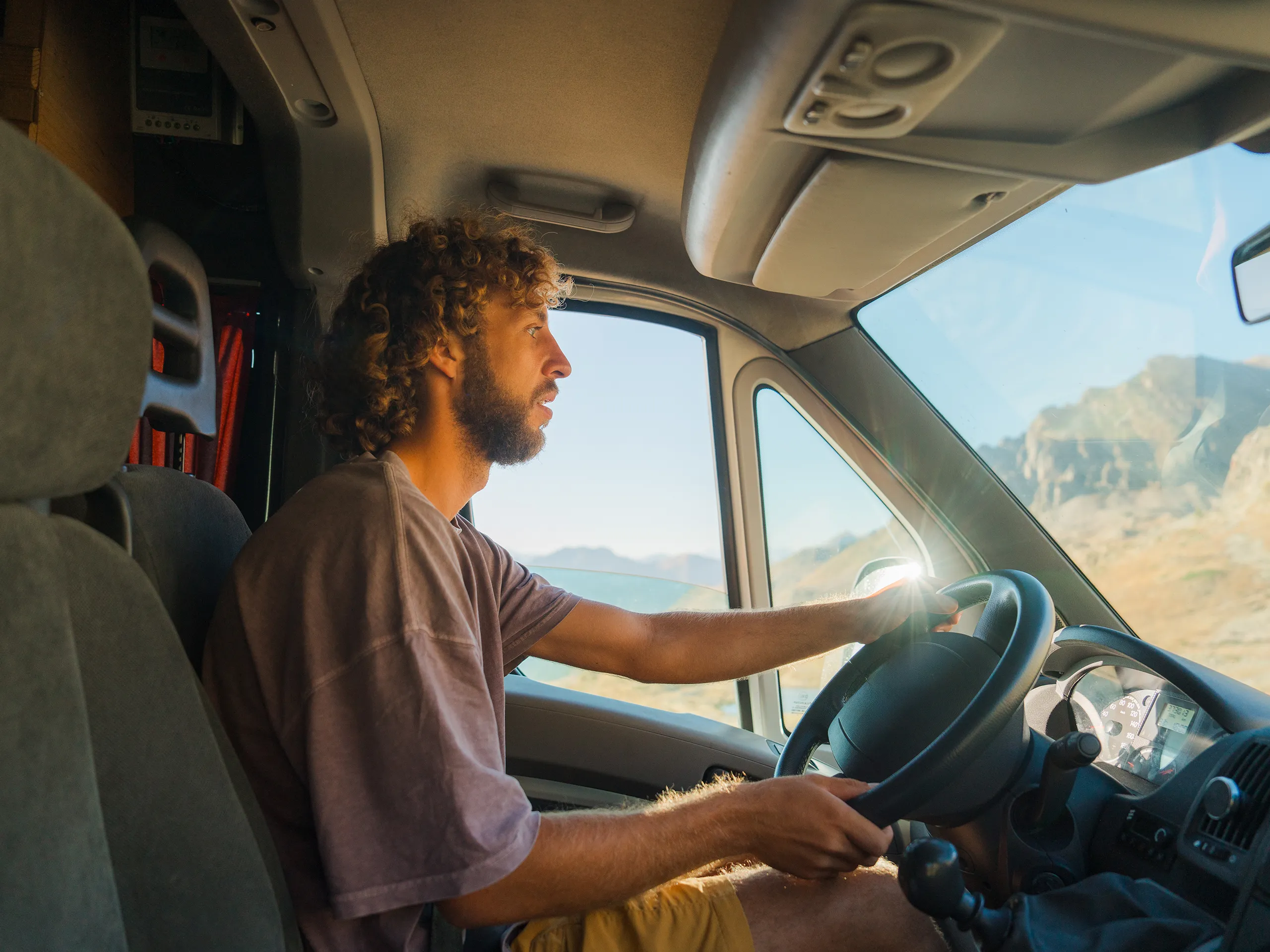 Man with curly hair drives a camper through a sunny mountain landscape.
