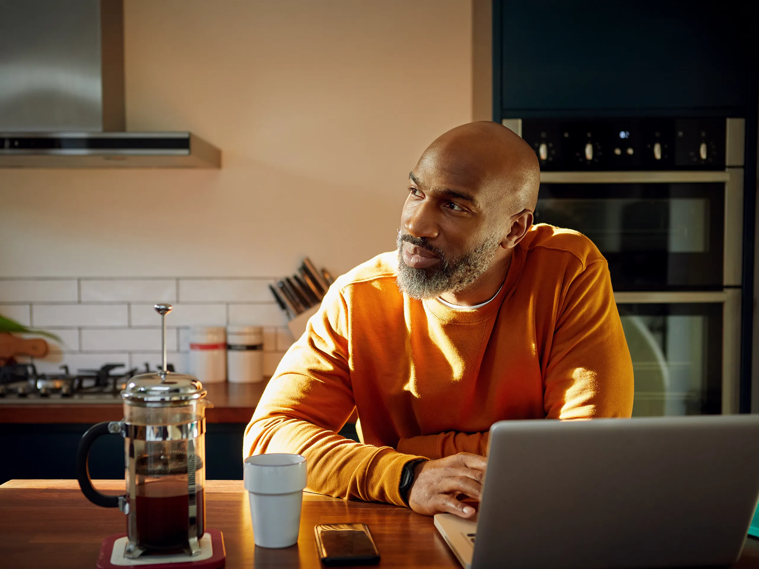 Man with a laptop sits thoughtfully in the kitchen with coffee.