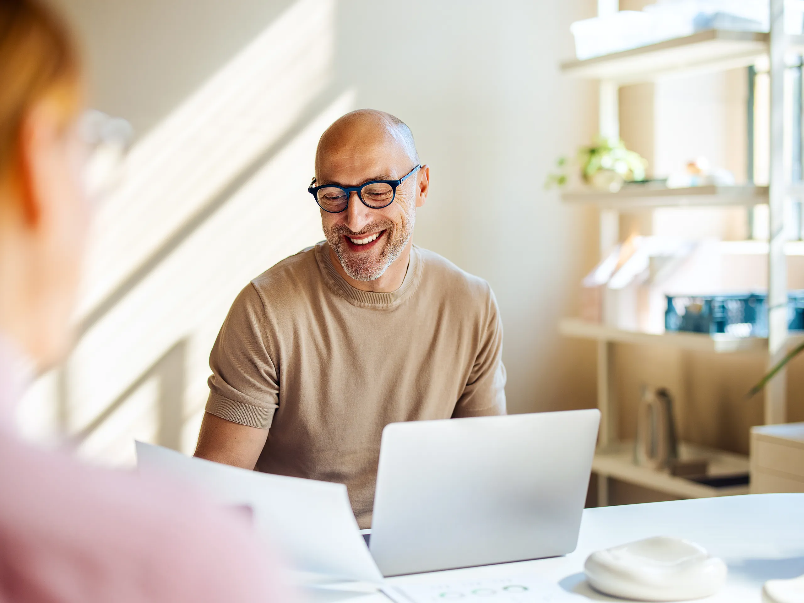 Smiling man with glasses working with a laptop and documents at a desk.