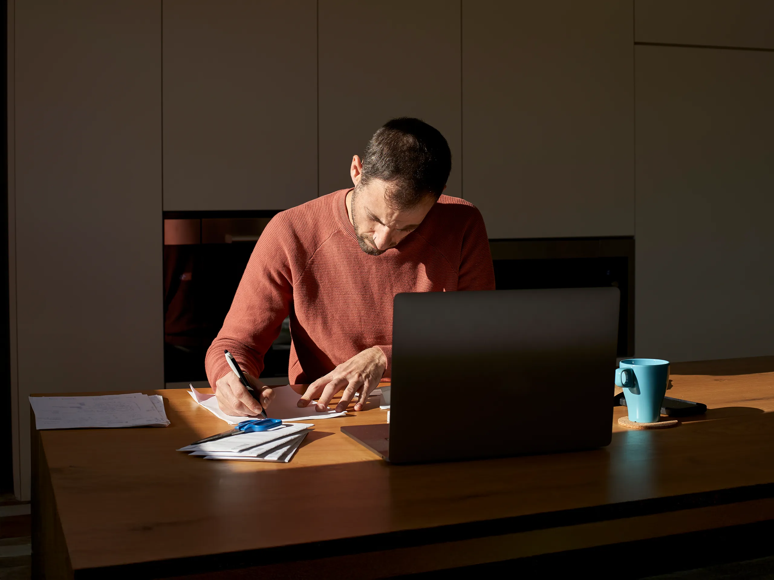 Man works with focus using a laptop and documents at a wooden table.