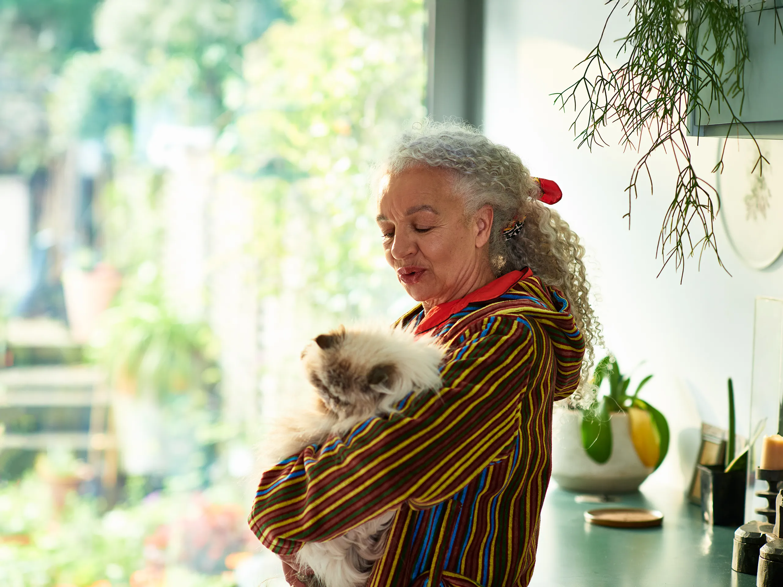 Older woman with curly hair holds a fluffy cat in a sunny kitchen.