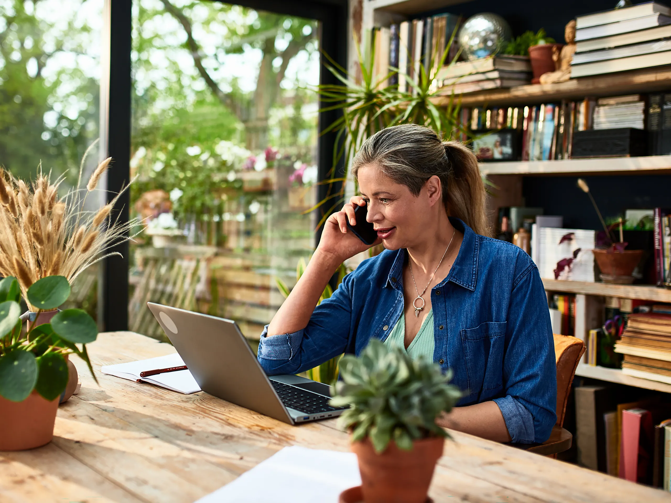 Woman talks on the phone while working on her laptop in a bright room with plants.