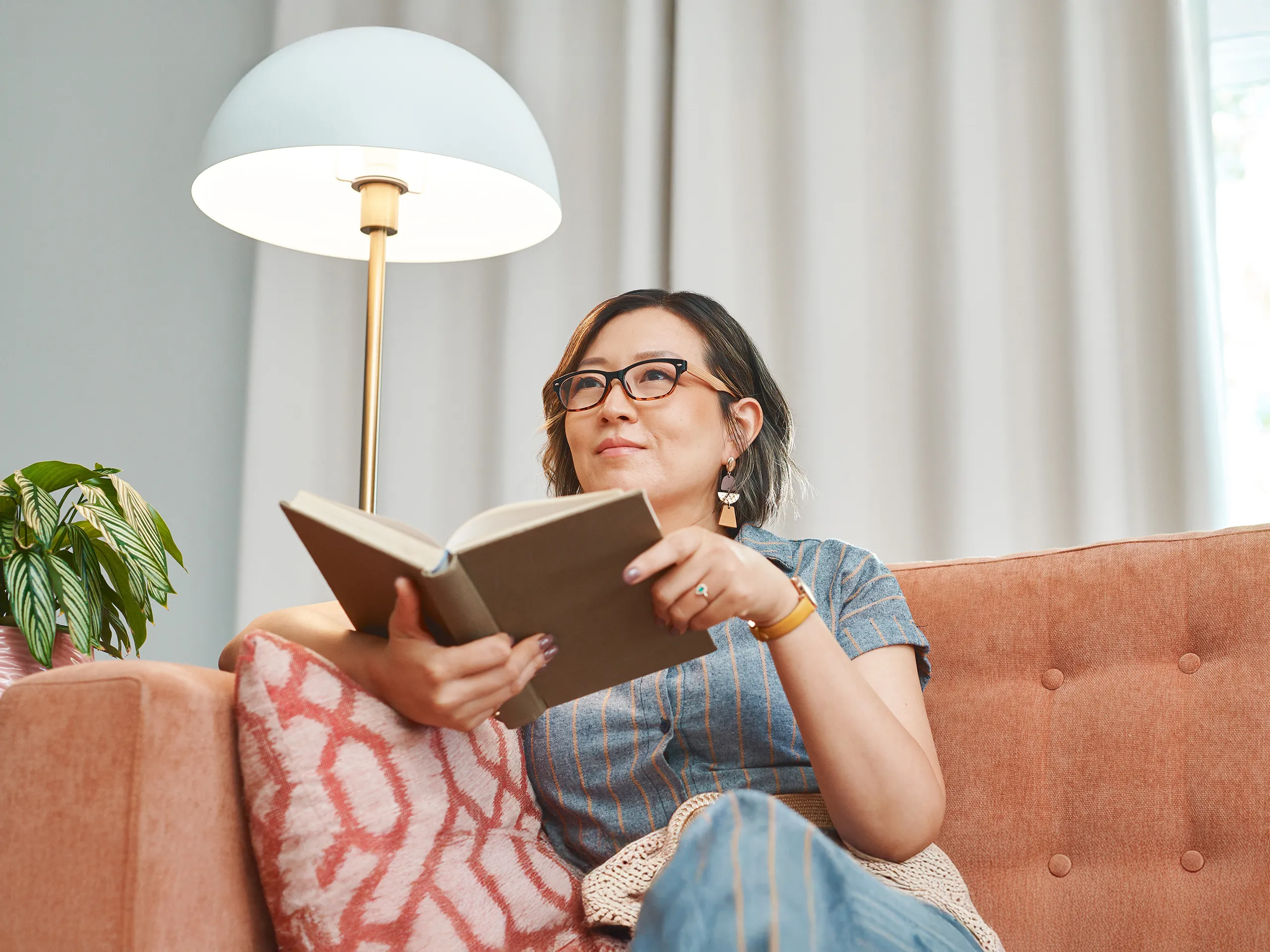 Woman reading a book on a sofa next to a floor lamp.