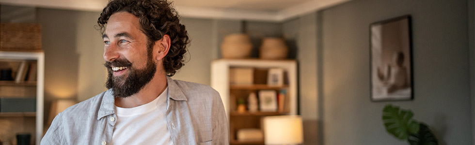 A middle aged man with a beard, white t shirt and shirt stands smiling in his home. The room he is in has soft warm homely tones.